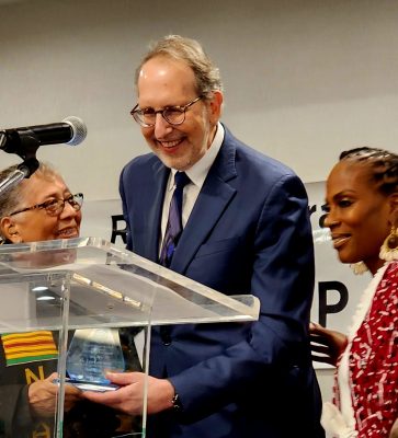 A man in a suit stands at a podium and is handed an award from a woman.