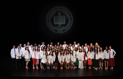A large group of medical school students dressed in their white coats stand on stage underneath the school seal. 